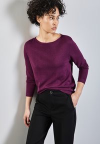 Lila gestrickter Pullover mit rundem Halsausschnitt und Dreiviertelärmeln, kombiniert mit schwarzen, taillierten Hosen mit Fronttaschen.