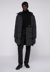 Abrigo de plumón negro con acolchado horizontal, combinado con un gorro de punto negro, bufanda, guantes y pantalones y zapatos negros ajustados.