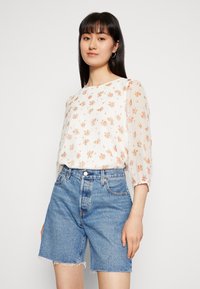 Blusa branca com pequenos padrões florais, mangas bufantes semi-transparentes; conjugada com calções de ganga azuis de cintura alta, com bainha desfiada.