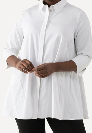 Person wearing a white collared, long-sleeve blouse with pleats, fastening buttons, paired with black pants on a plain background.