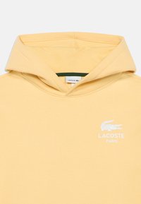 Κίτρινο φούτερ με κουκούλα από απαλό ύφασμα με τσέπη καγκουρό, διαθέτει λευκό λογότυπο Lacoste στο αριστερό στήθος και πράσινη εσωτερική επένδυση.
