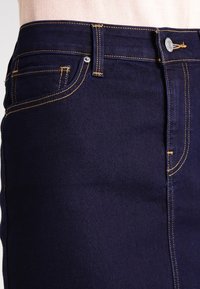 Mörkblå denimjeans har kontrasterande gula sömmar, en framficka och knappstängning. Slät textur med en åtsittande design.