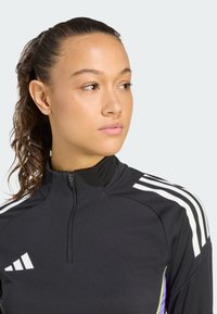Žena s skupljenom kovrčavom kosom nosi crni sportski duksericu na zip s bijelim prugama i gleda udesno na jednostavnoj pozadini.