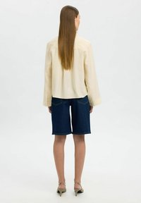 Camisa de manga larga color beige claro con un corte relajado, combinada con pantalones cortos de mezclilla azul oscura que llegan a medio muslo, y sandalias de tacón en color topo.