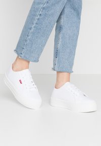 Baskets en toile blanche avec un bout arrondi, un design à lacets et une finition texturée. Elles disposent d'une semelle en caoutchouc blanche avec une légère plateforme.