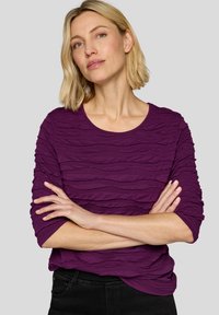 Lila Langarmshirt mit wellenförmigem Texturmuster, runder Ausschnitt und engen Ärmeln. Getragen mit schwarzen Jeans vor einem neutralen Hintergrund.