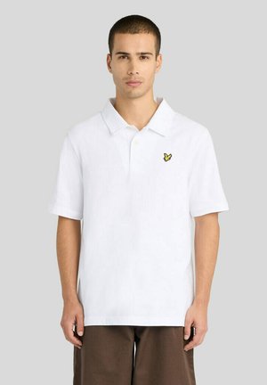 Man met kort haar, gekleed in een wit poloshirt met een klein geel vogel-logo en een bruine broek, staand tegen een effen lichte achtergrond.