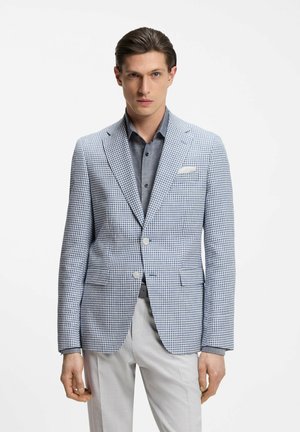 Man draagt een lichtblauw-wit pied-de-poule colbert over een grijs overhemd met knopen en witte broek, staand tegen een effen achtergrond.