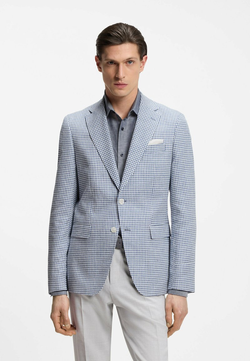 Uomo che indossa un blazer pied de poule azzurro chiaro e bianco sopra una camicia grigia con bottoni e pantaloni bianchi, in piedi davanti a uno sfondo semplice.