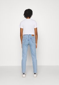 Persona con cabello rizado corto, vistiendo una camiseta blanca, jeans de cintura alta de color azul claro y zapatillas blancas, de pie frente a una pared blanca simple.