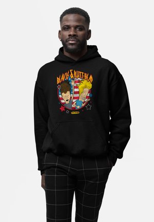 Hombre con sudadera con capucha negra con un gráfico colorido de Beavis y Butt-Head sobre un círculo con la bandera estadounidense, combinado con pantalones negros con patrón de cuadrícula blanca.