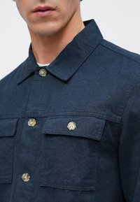 Laivastonsininen paita, jossa on jämäkkä kaulus, kaksi etutaskua ja pyöreät, vaaleat napit. Kangas on sileä ja hieman teksturoitu.