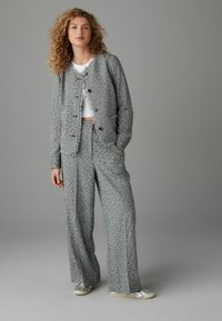 Dvoudílný outfit skládající se z texturované šedé bundy s roztrhanými okraji a odpovídajícími volnými kalhotami. Doplněno stříbrnými botami, na šedém pozadí.