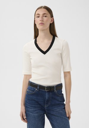Frau mit glatten braunen Haaren trägt ein weißes V-Ausschnitt-Top mit schwarzem Rand und blaue Jeans mit schwarzem Gürtel, stehend vor einem schlichten weißen Hintergrund.