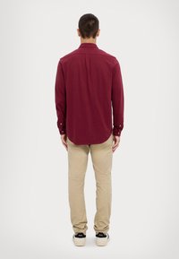 Camisa de manga larga color granate con puños de botones, dobladillo redondeado y textura suave, combinada con pantalones khaki claros y zapatillas negras.