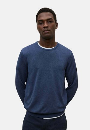 Hombre que lleva un jersey azul liso de manga larga sobre una camiseta blanca, de pie con las manos detrás de la espalda frente a un fondo liso.