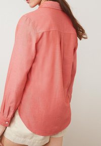 Chemise corail à manches longues avec boutons, en tissu doux et texturé. Elle présente un col classique, un pli simple au dos et un ourlet arrondi.