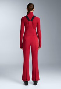 Piros overall teljes hosszúságú, bőszárú nadrággal, bordázott felsőrésszel és fekete vállpántokkal. A bokán cipzárdíszítéssel rendelkezik. Simább textúrájú.