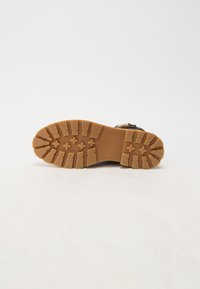Semelle en caoutchouc marron avec une surface texturée, dotée de profondes rainures et de motifs croisés. Logo circulaire au centre.