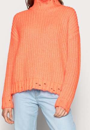 Femme portant un pull à col roulé en maille orange vif avec un ourlet usé et un jean bleu clair, debout devant un fond uni.