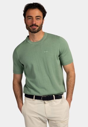 Man met donker haar en baard, draagt een groene trui met korte mouwen, beige broek en een zwarte riem, staat met handen in de zakken.