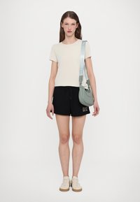 T-skjorte i lys beige, kombinert med svarte shorts med logo. Tilbehør inkluderer en grønn veske og beige sneakers.