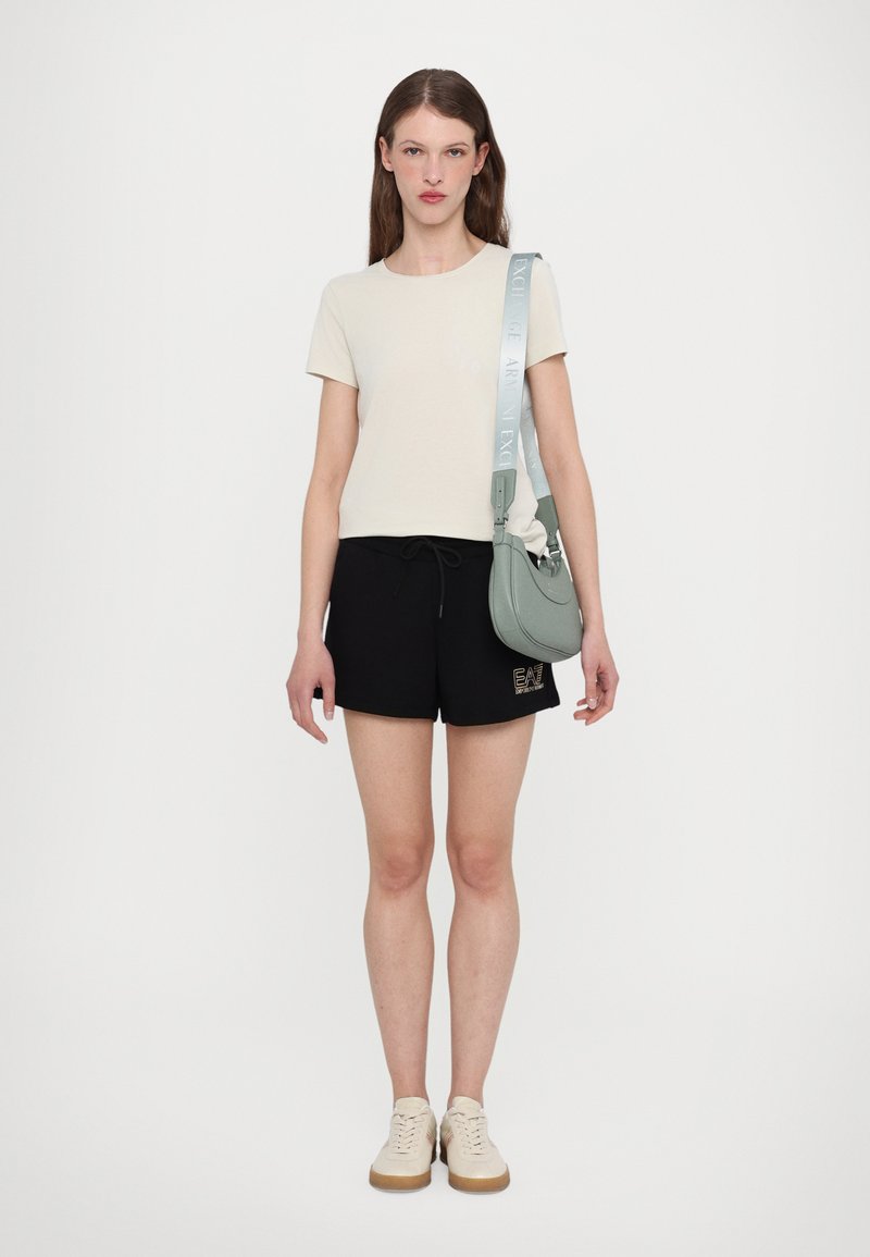 T-skjorte i lys beige, kombinert med svarte shorts med logo. Tilbehør inkluderer en grønn veske og beige sneakers.