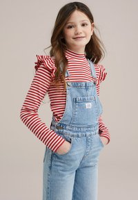 Pantalones de mezclilla en azul claro con tirantes ajustables, combinados con una blusa de rayas rojas y blancas de manga larga con mangas acampanadas.