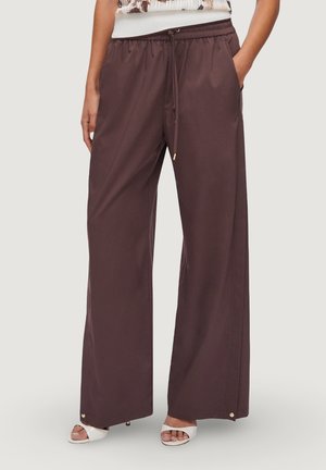 Donna che indossa pantaloni larghi marrone scuro con coulisse in vita e sandali bianchi aperti sulle dita, in piedi con le mani nelle tasche.