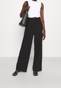 Schwarze, weit geschnittene Hose mit einem Gürtel in der Taille, kombiniert mit einem weißen ärmellosen Oberteil. Das Model trägt eine schwarze strukturierte Bucket-Bag und schwarze Sandalen.