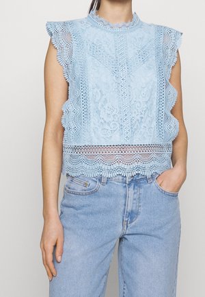 Ærmeløs lyseblå blonderbluse med bølget kant, båret med højtaljede blå denimjeans, med den ene hånd i lommen, foran en ensfarvet baggrund.