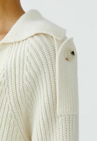 Nahaufnahme einer Schulter eines cremefarbenen Strickpullovers mit diagonaler Rippung, einem weiten Kragen und einem dekorativen Tab, der mit einem großen Knopf befestigt ist.