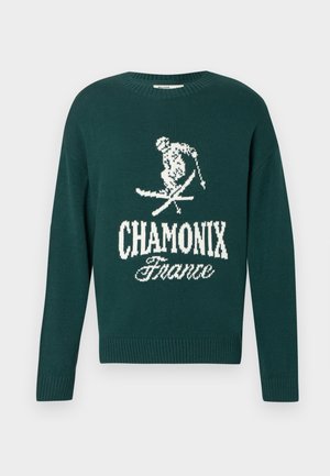 Zeleni pleteni pulover z belim grafičnim motivom smučarja in napisom "CHAMONIX Francija". Žepek in rob sta dopolnjena z rebrastim designom.