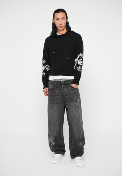 Pull noir avec motifs de crâne et d'os blancs sur les manches, associé à un pantalon en jean gris présentant une texture usée et des détails de logo.