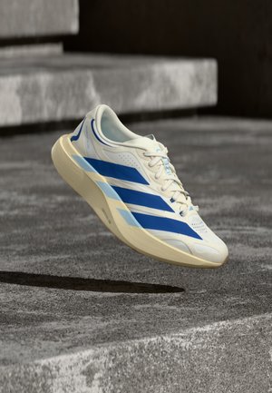 Chaussure de course Adidas blanche et bleue suspendue au-dessus d'une marche en béton avec un fond de surface rugueuse texturée.