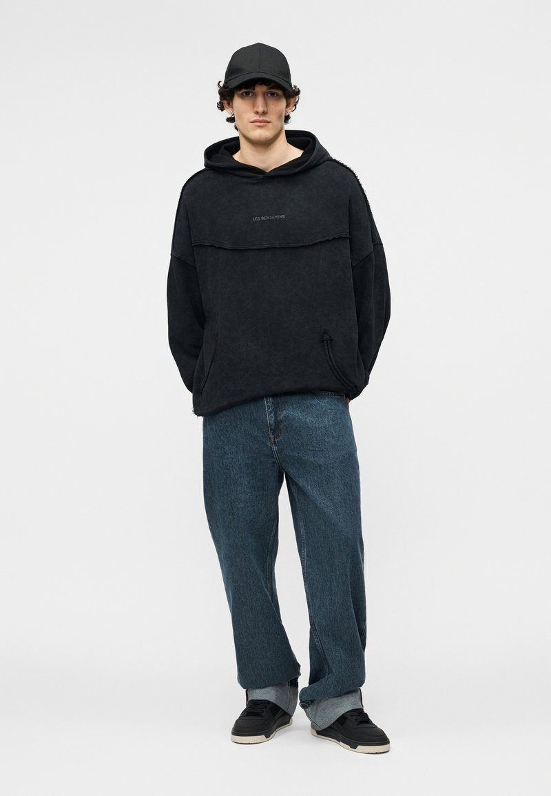 Junger Mann trägt einen schwarzen Hoodie, eine schwarze Kappe, lockere blaue Jeans mit Umschlägen und schwarze Sneakers, steht vor einem schlichten hellen Hintergrund.