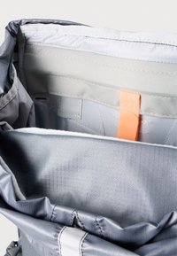 Interiér šedého látkového batohu s hladkým podšívkou, oranžovým štítkem, mřížkovaným vzorem a bočními kapsami. Obsahuje bezpečné uzavírání a nastavitelné popruhy.