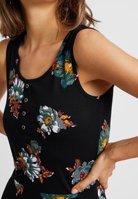 Černý top bez rukávů s květinovými vzory v tónech modrozelené, bílé a oranžové. Vybaven kulatým výstřihem a knoflíkovými detaily vpředu. Měkká textilie.