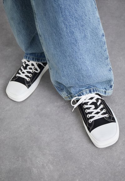 Svarta canvas-sneakers med vita gummitådelar och snören, i kombination med ljusblå baggyjeans. Grå texturerad yta nedanför.