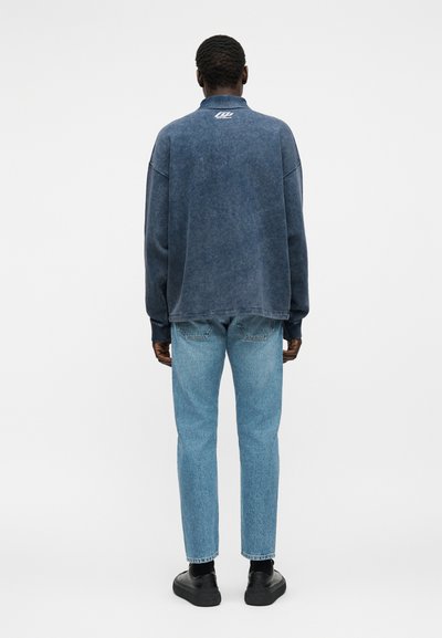 Άνδρας που φοράει ξεθωριασμένο μπλε πουλόβερ με λευκό λογότυπο στον λαιμό και ανοιχτό μπλε τζιν, στέκεται αντικρίζοντας ένα απλό ανοιχτό γκρίζο φόντο.