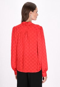 Rød bluse med lange ærmer, prikmønster og en plisseret rygdesign. Stoffet ser glat ud med en let gennemsigtig tekstur.