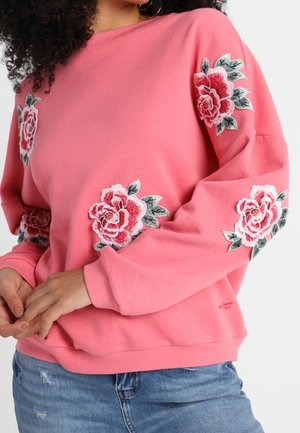 Femme portant un sweat-shirt rose avec des roses roses et blanches brodées sur les manches et le devant, associé à un jean bleu.