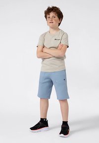 Chlapec s kudrnatými červenými vlasy stojí s překříženýma rukama, na sobě má béžové tričko Champion, modré kraťasy, černé ponožky a černé tenisky na bílém pozadí.