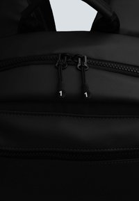Zaino nero con chiusure a zip doppie dotate di tirazip numerici, texture della stoffa liscia e una tasca frontale con zip per ulteriore spazio di archiviazione.