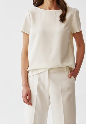 Kobieta ubrana w kremową bluzkę z krótkim rękawem wpuszczoną w kremowe, plisowane spodnie z wysokim stanem, z jedną ręką w kieszeni.