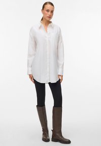 Hvit oversized skjorte med knapper og lomme, brukt over svarte leggings og brune knelengde støvler. Glatt stoff, klassisk krage, lange ermer.