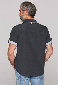 Donkergrijs shirt met korte mouwen en opgerolde manchetten, gestreepte details op de binnenmouwen en een kleine rood-wit-blauwe accent aan de achterkant van de kraag.