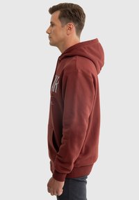 Bordeauxfarbener Kapuzenpullover mit einer Fronttasche, Kordelzugkapuze und grafischem Text. Hergestellt aus weichem Baumwoll-Mischgewebe, mit gerippten Bündchen und Saum.