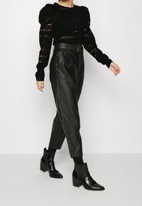 Pull noir transparent avec manches bouffantes, associé à un pantalon en cuir noir taille haute et des bottines noires brillantes à finition texturée.