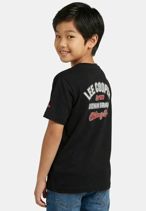 Garçon souriant portant un T-shirt noir Lee Cooper et un jean bleu, légèrement tourné pour montrer le texte au dos du T-shirt.